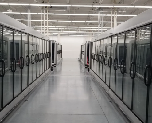 20180515_113124-1920w A wide aisle lined with glass-fronted refrigeration units in a well-lit store, showcasing empty spaces for cooling products.
