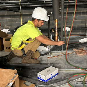 A worker in a yellow shirt and white hard hat kneels on a floor surrounded by tools and boxes, focusing on pipe installation in a construction area.