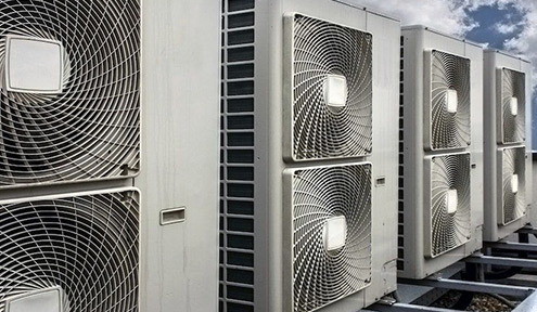 Row of industrial air conditioning units mounted on a building rooftop against a backdrop of blue sky and scattered clouds.
