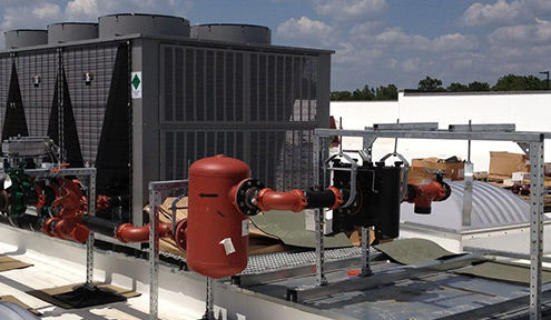 Industrial rooftop HVAC system with large cooling units, pipes, and valves set against a blue sky and white building.
