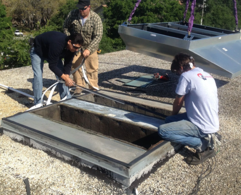 Workers are installing a large metal component on a roof, surrounded by gravel and green trees under a clear blue sky.