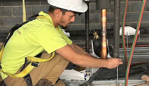 A worker in a bright yellow shirt and harness kneels on a construction site, adjusting piping among tools and boxes.