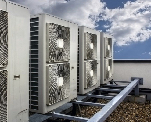 Row of industrial air conditioning units mounted on a building rooftop against a backdrop of blue sky and scattered clouds.